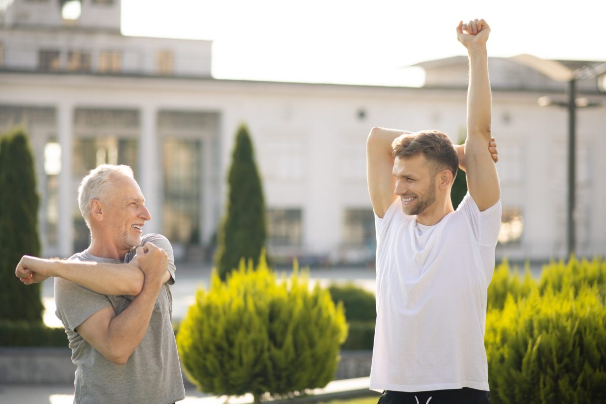 Morning workout. Young bearded male and mature grey-haired male stretching arms outside, looking at each other, smiling